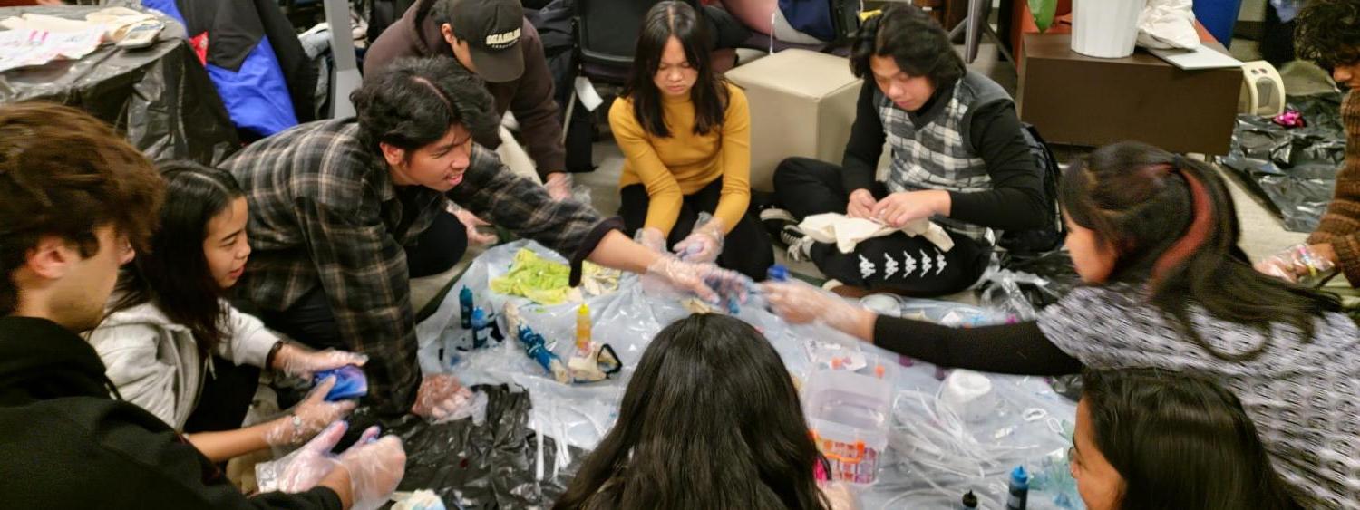 Students sitting in circle on floor tie dying tote bags with group of students around table knitting in background