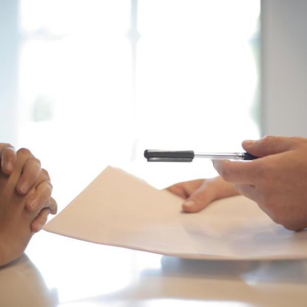 a person in a suit hands a paper and pen so the person sitting at the desk with them for their signature