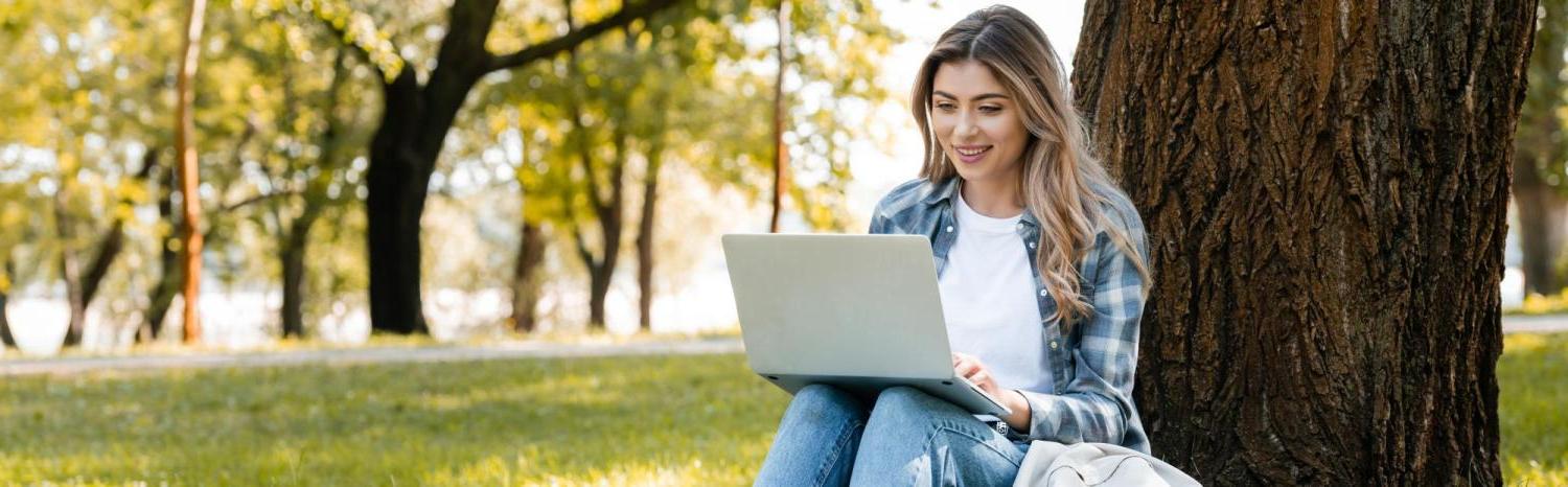 Photo of a student on a laptop outside under a tree on a sunny day.