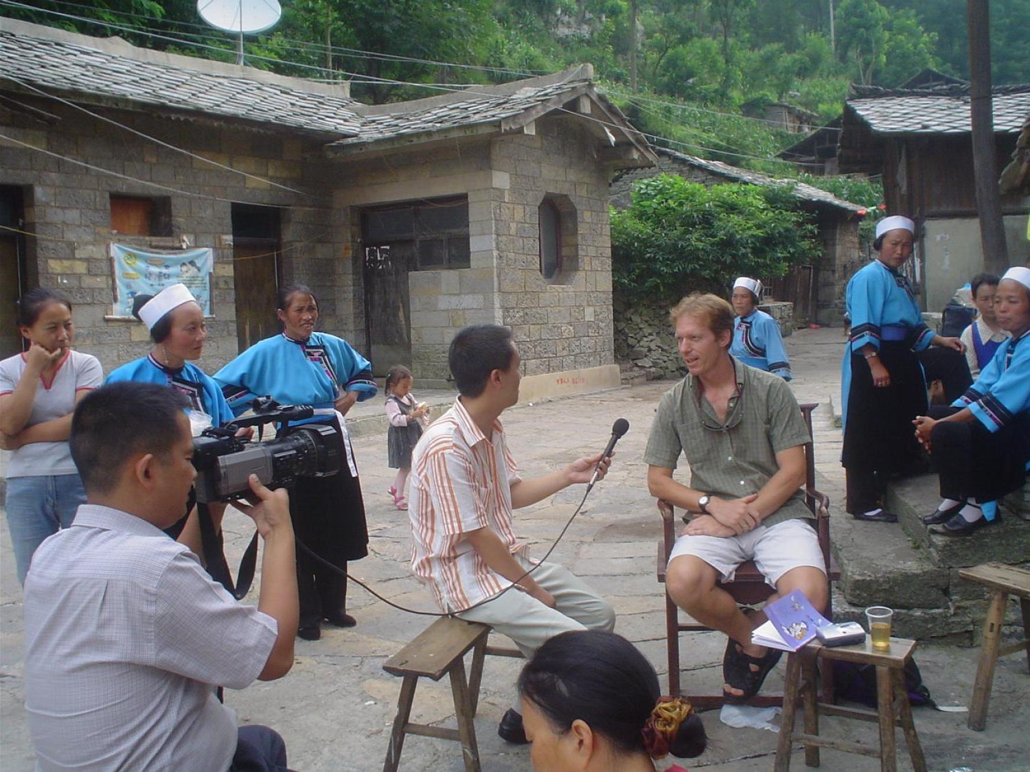 在中国农村，一名白人男子坐在电视采访者和摄制组旁边，村民们在一旁观察