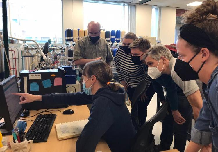 坐在电脑前, Sylvia demonstrates part of the Stable Isotope measurement process to a group of Finnish visitors closely gathered around her