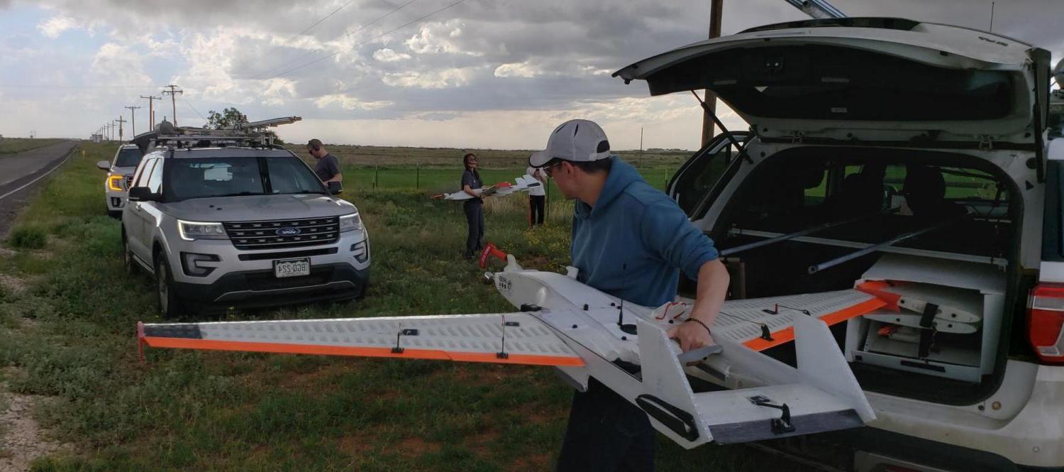 Man lifts drone with fixed wings out of the trunk of an SUV while other scientists work in background, and storm clouds brew above