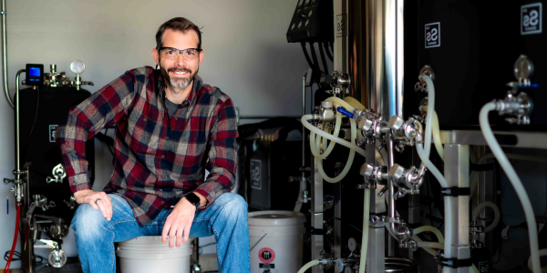 Man in flannel shirt wearing safety glasses sits next to equipment with lots of tubes