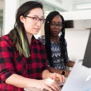 Two women working on a laptop computer at a standing desk. 