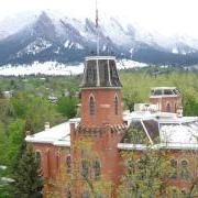 The Old Main building surrounded by the green foliage of spring trees, with snow-dusted foothills in the background.