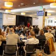 Numerous people sit at large round tables in a conference space, turning their attention to a speaker holding a microphone and presenting from a lecturn. 